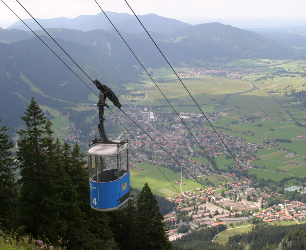 Seilbahn fährt einen Berg hinauf, mit einer kleinen Stadt unten sichtbar, umgeben von Grünflächen und Hügeln unter einem klaren blauen Himmel.