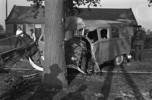 Schwarz-weißes Bild eines verunglückten Autos am Straßenrand mit einem Baum im Vordergrund, einigen Menschen in der Nähe und Häusern, Bäumen und einem Mast im Hintergrund.