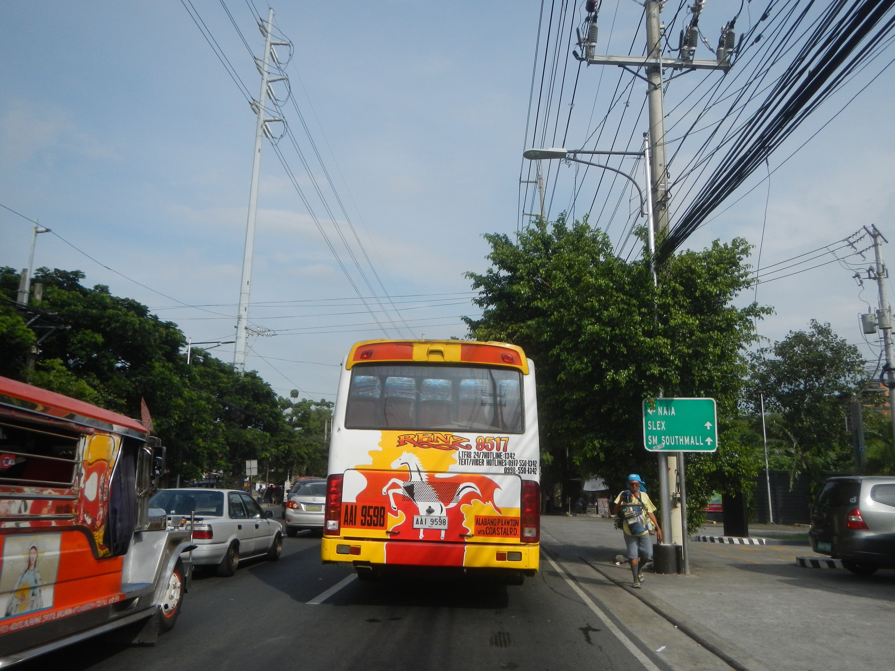 Ein gelb-roter Bus fährt auf einer Straße mit hohen Strommasten und -leitungen, eine Person geht auf dem Gehweg, ein grünes Schild ist sichtbar, Bäume und ein klarer blauer Himmel sind im Hintergrund.