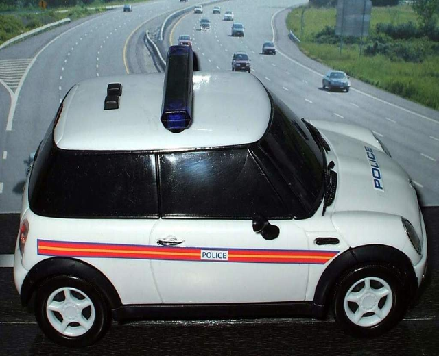 Weißes Polizeiauto mit 'Polizei'-Aufschrift auf einer Straße außerhalb einer Stadt, umgeben von Verkehr und gesäumt von Gras und Bäumen.