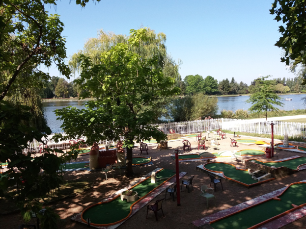 Minigolf-Platz in einem Park mit grünem Gras, Bäumen und einem Zaun, umgeben von verstreuten Stühlen und Tischen, einem Gewässer mit einem Boot und sichtbarem Himmel im Hintergrund.