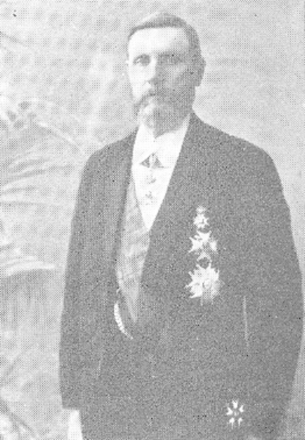 Ein Mann in einem schwarzen und weißen Anzug mit einem weißen Hemd und schwarzer Krawatte steht vor einer üppigen grünen Pflanze, sein Gesichtsausdruck ernst.