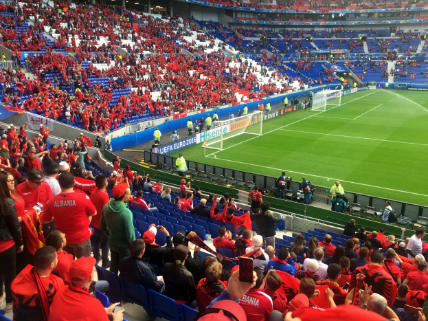 Ein volles Stadion mit sitzenden und stehenden Zuschauern, Fahnen, einem Tor und Deckenleuchten, während die Menge ungeduldig auf den Beginn eines Fussballspiels wartet.