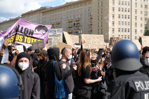 Gruppe maskierter Demonstranten mit Schildern vor einem Gebäude mit zwei Polizisten in Helmen, Bäumen und bewölktem Himmel im Hintergrund.