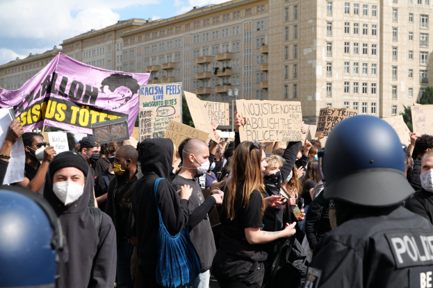 Gruppe maskierter Demonstranten mit Schildern vor einem Gebäude mit zwei Polizisten in Helmen, Bäumen und bewölktem Himmel im Hintergrund.