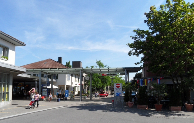 Eine Stadtstraße mit Fußgängern, einige mit Kinderwagen, Gebäuden, Bäumen, Topfpflanzen, Schildern, Fahrzeugen und einem bewölkten Himmel.