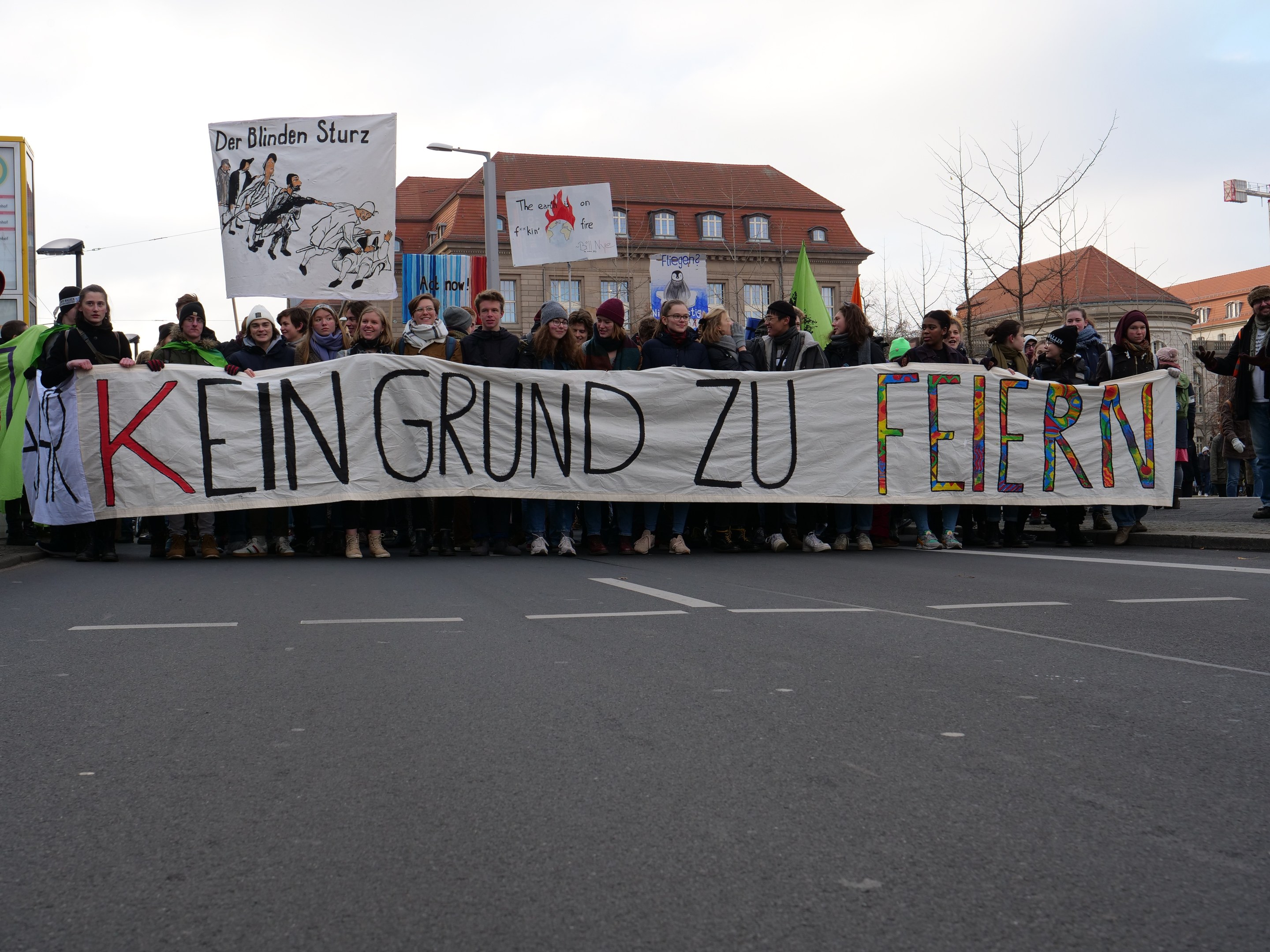 Demonstranten halten ein Transparent mit der Aufschrift "Kein Grund zu Feiern" gegen die deutschen Sparmaßnahmen, mit Gebäuden, Bäumen und einem klaren Himmel im Hintergrund.