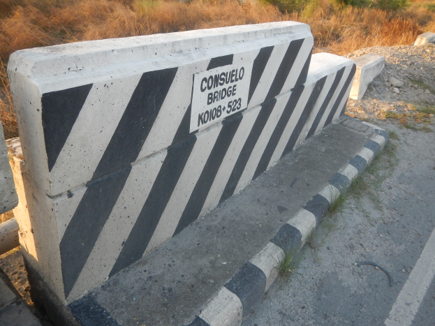 Schwarz-weiß gestreifter Betonbarriere mit Text an einer Straßenrand, mit Vegetation im Hintergrund.
