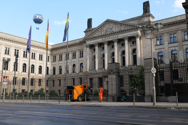 Großes modernes Regierungsgebäude mit vielen Fenstern, umgeben von Straßeninfrastruktur, Fußgängern und Bäumen, mit einem bewölkten Himmel im Hintergrund.