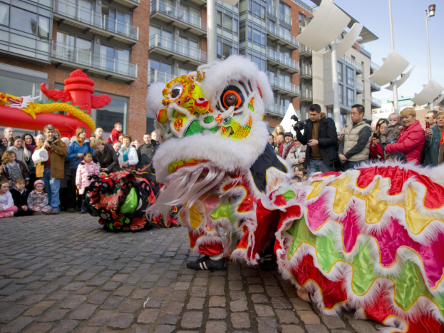 Ein lebendiges chinesisches Neujahrsfest in Amsterdam mit einer Löwen-Tanzvorführung im Vordergrund mit einer Zuschauermenge, einige halten Kameras, vor einem Hintergrund aus Gebäuden, Laternenmasten und einem klaren blauen Himmel.