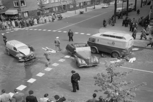 Schwarz-weiß-Szene eines Autounfalls an einer Straßenkreuzung mit mehreren Fahrzeugen, darunter ein Van, und einer Gruppe von Zuschauern; Gebäude, Bäume, Strommasten und Texttafeln im Hintergrund.