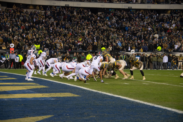 Ein Fußballspiel im Stadion mit Spielern in Sportbekleidung und Helmen auf dem Rasen, umgeben von Zuschauern und Stadionelementen im Hintergrund.