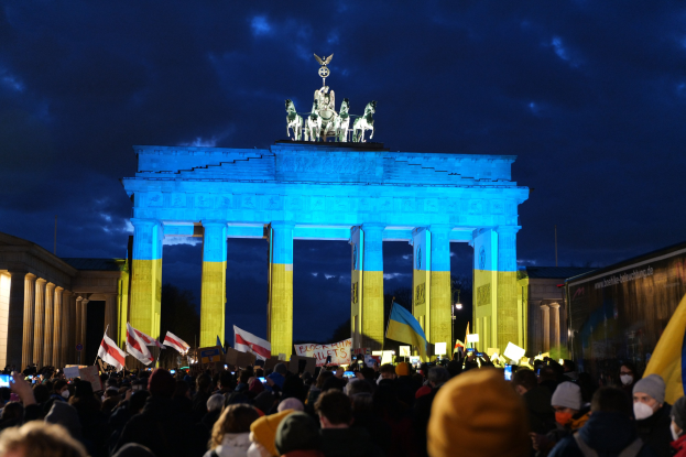 Eine Menschenmenge steht vor dem Brandenburger Tor in Berlin, Deutschland, mit Fahnen und Plakaten in den Händen, einem Banner auf der rechten Seite und dem Tor mit Statuen und Säulen geschmückt, unter einem bewölkten Himmel.