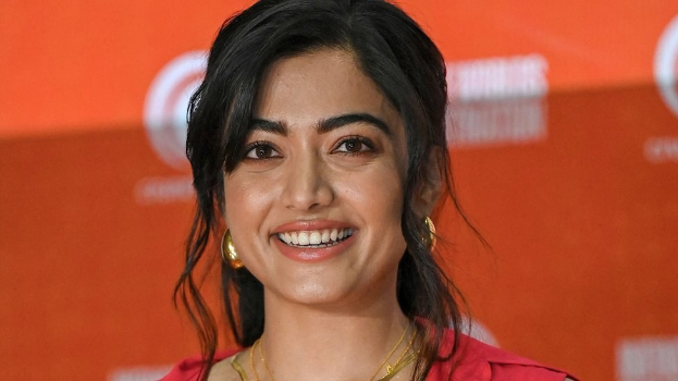 Eine Frau in einem roten Kleid mit einem eleganten Dutt und dezentem Make-up lächelt in die Kamera, trägt schwarze High Heels und hat einen hellgelben Banner im Hintergrund mit der Aufschrift "Sonam Kapoors beste Beauty-Looks vom Cannes Film Festival".