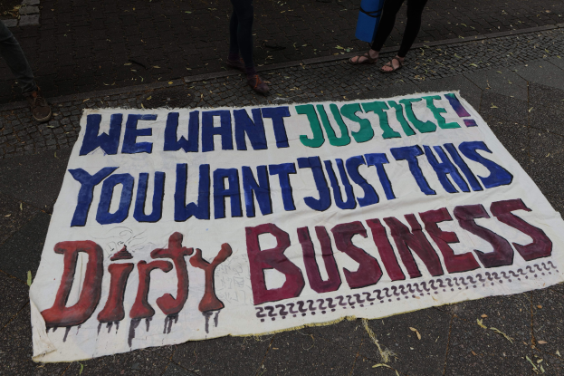 Banner an einer Straße mit dem Text "Wir wollen Gerechtigkeit, ihr wollt nur diesen schmutzigen Deal" und zwei Personen obenstehend, eine hält ein blaues Objekt.