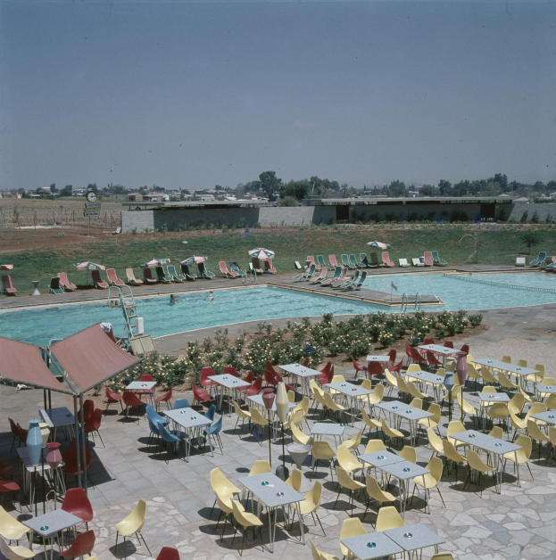 Freibad mit Tischen, Stühlen, Sonnenschirmen und umgebender Vegetation, umgeben von einem Zaun und einer Wand, unter einem bewölkten Himmel.