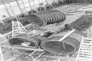 Ein Schwarz-Weiß-Foto eines Fabrikinnenraums mit einem großen zylindrischen Objekt in der Mitte, umgeben von Leitern, Stangen und anderen Gegenständen, mit einer Wand aus Fenstern im Hintergrund.