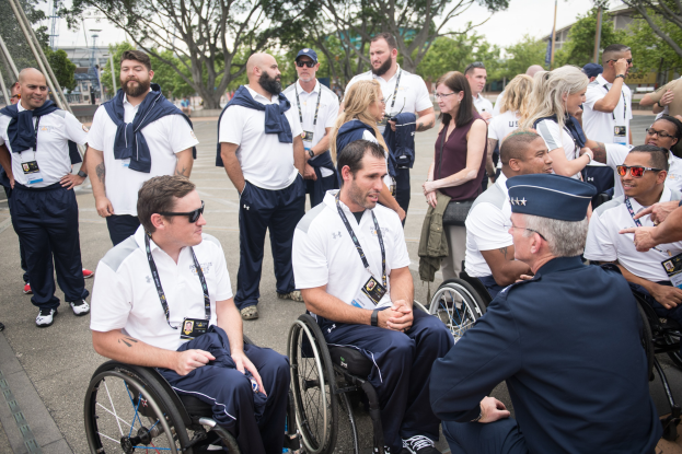Gruppe von Menschen im Rollstuhl, die im Freien miteinander sprechen, vor einem Hintergrund aus Bäumen, Gebäuden und einem klaren blauen Himmel.
