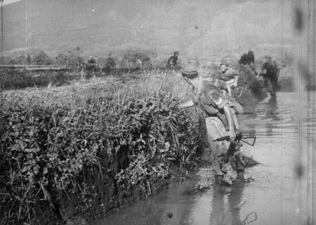 Schwarz-weißes Bild einer Gruppe von Männern, die durch einen Fluss gehen, einer hält eine Waffe, mit Pflanzen auf der linken Flussseite und Hügeln im Hintergrund unter einem klaren Himmel.