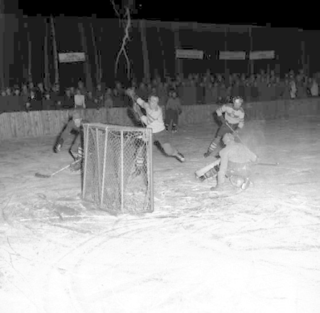 Schwarzes und weißes Foto eines Hockey-Spiels auf einem Eisstadion, mit Spielern in der Nähe eines Tors und Zuschauern im Hintergrund.