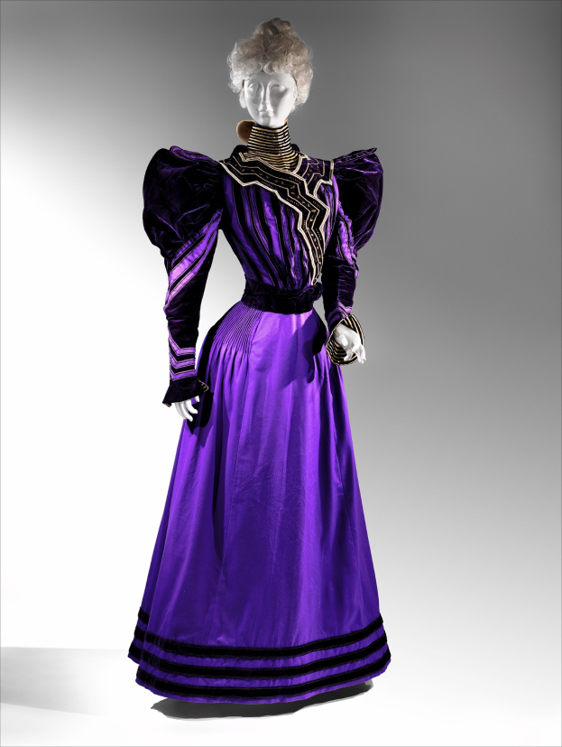 Eine Schaufensterpuppe in einem lila Abendkleid mit schwarzem und goldenem Muster, mit einer eng anliegenden Taille, einem langen bodenlangen Rock mit goldener Stickerei und Perlen sowie einer Spitzenborte an der Taille, kombiniert mit einem eleganten Hochsteckdutt und zarten Ohrringen.