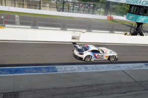 Ein Porsche 911 GT3 RSR fährt auf einer Rennstrecke mit einer Umrandung, Gras, Bäumen, Gebäuden und einem klaren blauen Himmel im Hintergrund, mit einer Tafel auf der rechten Seite, die die Teilnahme an den 24 Stunden von Le Mans anzeigt.