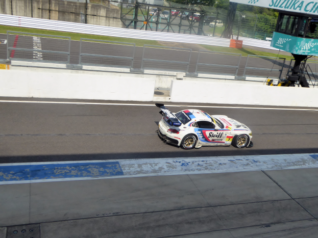 Ein Porsche 911 GT3 RSR fährt auf einer Rennstrecke mit einer Umrandung, Gras, Bäumen, Gebäuden und einem klaren blauen Himmel im Hintergrund, mit einer Tafel auf der rechten Seite, die die Teilnahme an den 24 Stunden von Le Mans anzeigt.