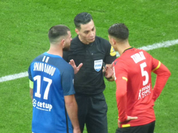 Schiedsrichter in schwarzem Hemd und Hose, der mit zwei Männern in Brille auf einem Fußballfeld spricht, einer trägt ein blau-rotes T-Shirt mit Text.