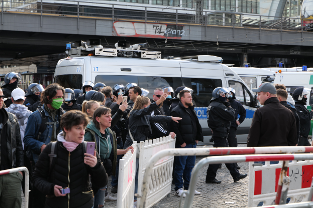Eine Gruppe von Menschen steht vor Polizeifahrzeugen, einige tragen Helme und halten Telefone, mit Absperrungen im Vordergrund und einer Brücke mit Geländern und Gebäuden im Hintergrund in Berlin, Deutschland.