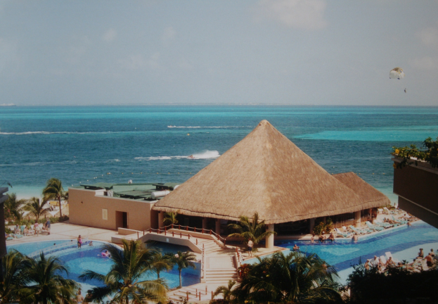 Resort mit einem gro├čen Schwimmbad umgeben von Palmen, Menschen im Wasser, einem Ozean im Hintergrund, einer bew├Âlktem Himmel und einem Fallschirm über dem Himmel.