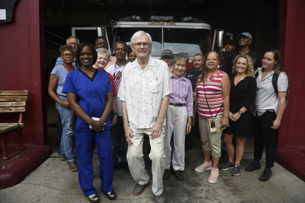 Eine Gruppe von Freiwilligen vor einem Feuerwehrauto, mit einer Bank links und einer Wand im Hintergrund.