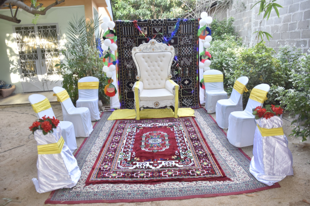 Hochzeitszeremonie in einem Garten mit weißen Stühlen, einem roten Teppich, Blumenbouquets, Dekorationsgegenständen, Ballons, Pflanzen, Bäumen, einem Gebäude mit Fenstern und einer Tür und einer Wand auf der rechten Seite.