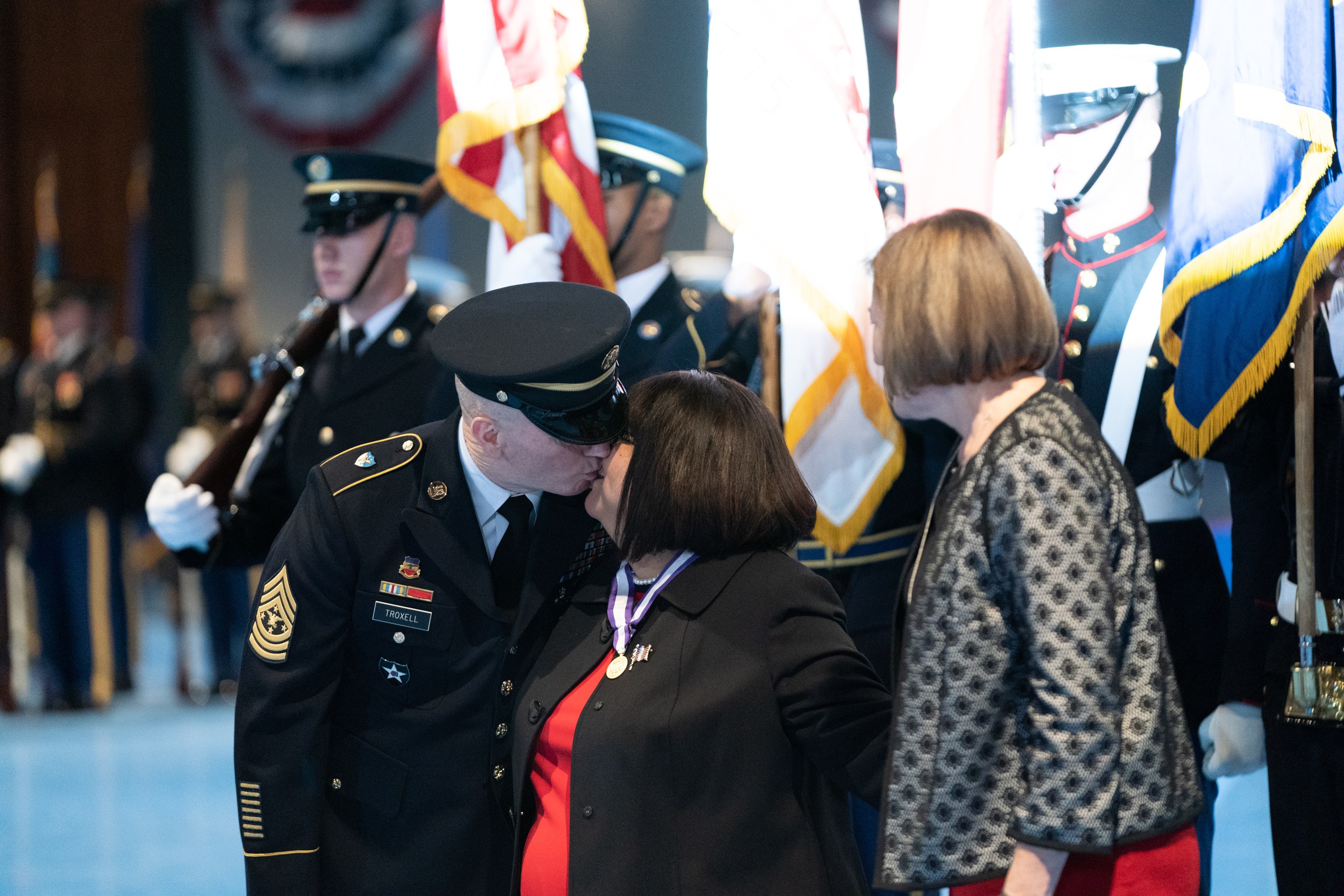 Ein Mann und eine Frau küssen sich, während sie sich in Gegenwart einer Gruppe von Menschen in Uniform und Mützen umarmen, einige halten Fahnen.