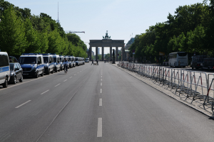 Lange Reihe von Polizeiwagen, die auf der Straße vor dem Brandenburger Tor in Berlin geparkt sind, mit Menschen auf Fahrrädern und Barrieren, Bäumen an den Seiten und einem Bogen mit Statuen im Hintergrund.