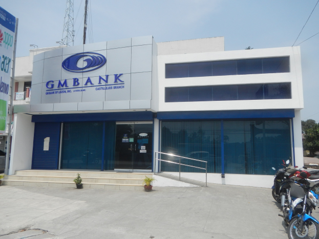Außenansicht der Gmbank Bank in Hyderabad mit Glaswänden, Treppen mit Geländern, Topfpflanzen, Straße mit Fahrzeugen, Banner, Versorgungsmast, Bäumen und bewölktem Himmel.