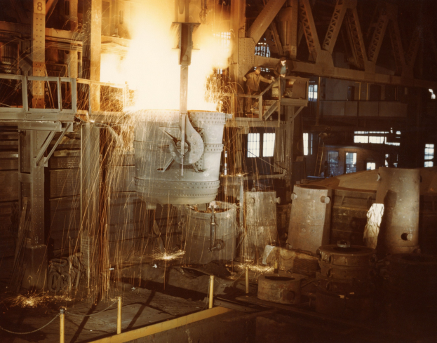 Großes Metallobjekt wird in einen Ofen in einer industriellen Umgebung gegossen, Arbeiter in der Nähe und durch das Feuer des Ofens beleuchtet, Metallobjekte, Pfosten und Fenster im Hintergrund.