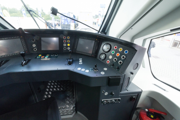 Cockpit eines Zuges mit Feuerlöscher im Vordergrund, das eine Glasfensteransicht von Menschen, Fahrzeugen, Bäumen, Masten, Planken und anderen Gegenständen außerhalb zeigt.