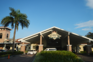 Der Eingang eines Resorts mit Palmen vorne, umgeben von Gebäuden, Fahrzeugen auf der Straße, Verkehrskegeln, Büschen, Bäumen und einem bewölkten Himmel im Hintergrund.