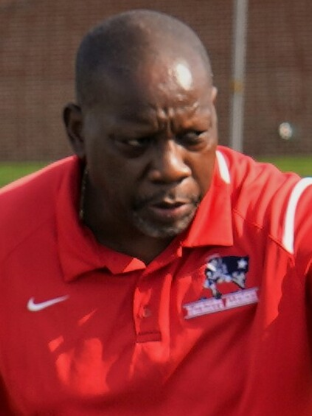 Ein Mann in einem roten T-Shirt steht auf einem Feld und hat die Hände in die Hüften gestützt, er sieht ernst aus, mit einem unscharfen Hintergrund.