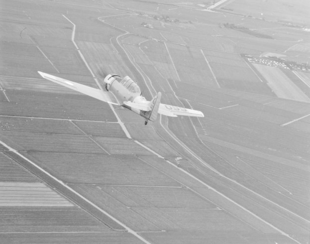 Schwarzes und weißes Foto eines kleinen Flugzeugs, das über ein Feld mit Kulturpflanzen fliegt und in der Mitte des Bildes zu sehen ist.