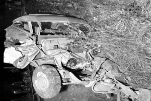 Ein Schwarz-Weiß-Foto eines schwer beschädigten Autos am Straßenrand, mit eingedellter Front, zersplitterten Fenstern, einem Baum im Hintergrund und verstreuten Gegenständen rund um die Unfallstelle.