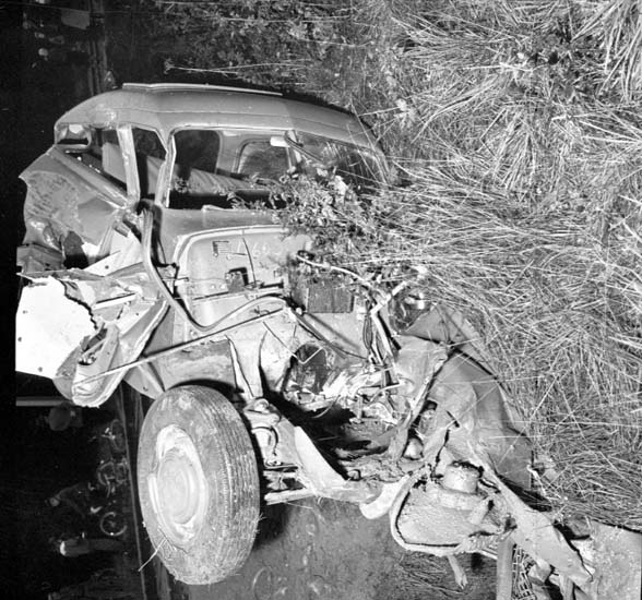 Ein Schwarz-Weiß-Foto eines schwer beschädigten Autos am Straßenrand, mit eingedellter Front, zersplitterten Fenstern, einem Baum im Hintergrund und verstreuten Gegenständen rund um die Unfallstelle.