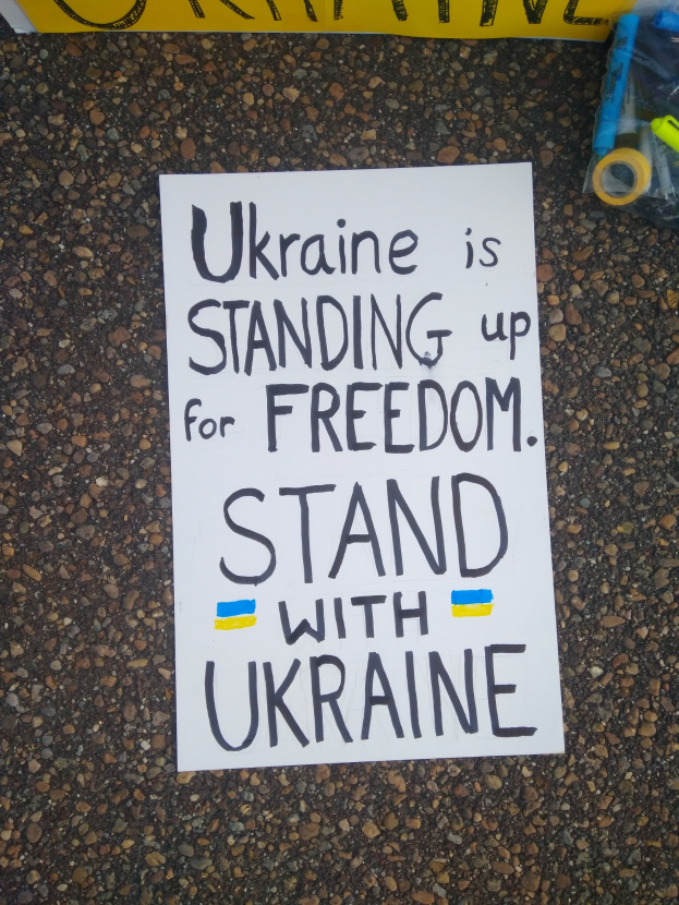 Ein weißer Zettel mit den Worten "Ukraine steht für Freiheit, steht mit Ukraine" liegt vor einer gelben Fahne auf dem Boden. Rechts davon befindet sich eine Plastikhülle mit verschiedenen Gegenständen.