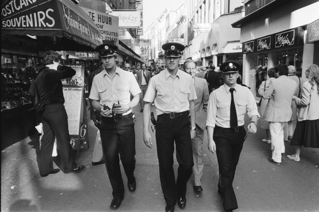 Drei Polizisten in Uniform gehen eine belebte Stadtstraße entlang, gesäumt von hohen Gebäuden, Geschäften und Passanten.