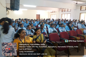 Eine Gruppe von Menschen sitzt in Stühlen in einem Raum mit einer Wand, Fenstern auf der rechten Seite, Deckenleuchten und einem Ventilator und einem Redner auf der linken Seite, mit Text und einer Karte unten.