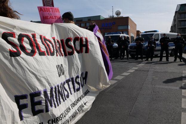 Gruppe von Menschen marschiert mit einem 'Solidarität und Feminismus'-Schild die Straße entlang, mit parkenden Fahrzeugen, Gebäuden, einer Schüsselantenne und einem klaren blauen Himmel im Hintergrund.