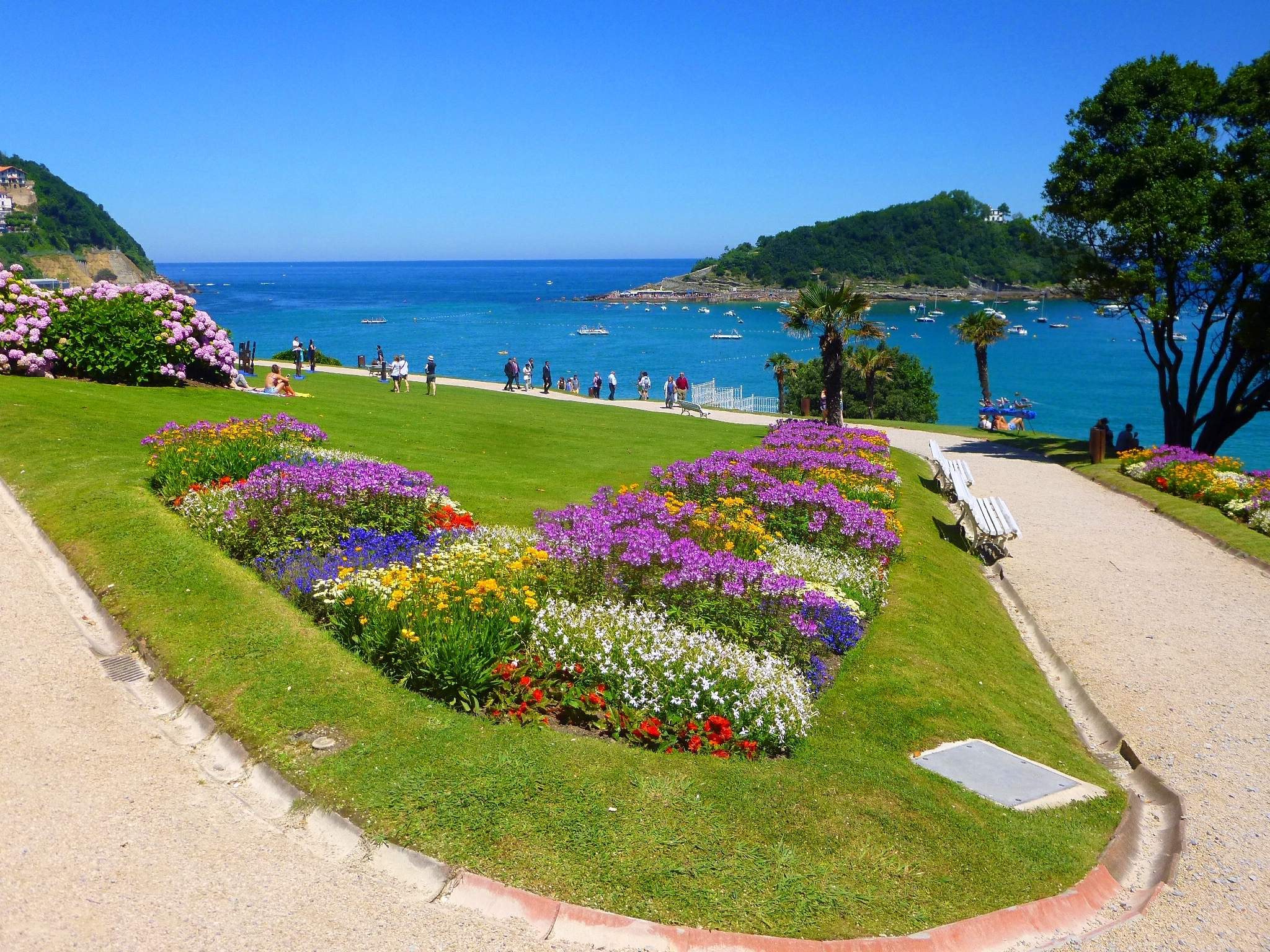 Ein Park mit bunten Blumen umgeben von grünem Gras und Bäumen, mit Bänken, Fußwegen, Menschen, Booten auf dem Wasser, Gebäuden und einem klaren blauen Himmel im Hintergrund.