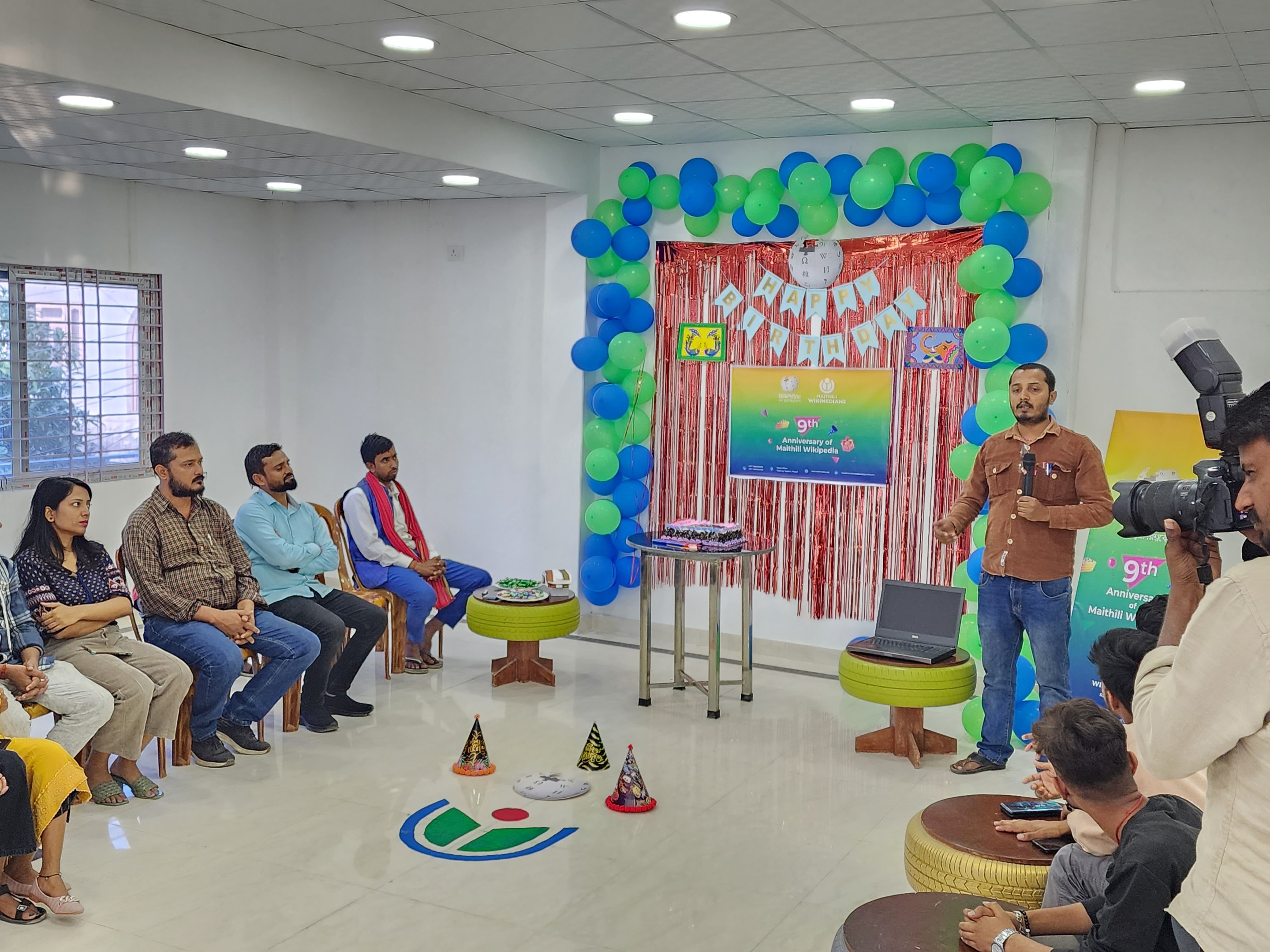 Eine Gruppe von Menschen sitzt auf Stühlen und schaut in eine Menschenmenge während einer 9. Geburtstagsfeier, wobei eine Person ein Mikrofon, ein Laptop, eine Tafel, Ballons und Deckenlampen hält.