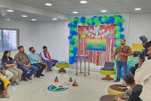 Eine Gruppe von Menschen sitzt auf Stühlen und schaut in eine Menschenmenge während einer 9. Geburtstagsfeier, wobei eine Person ein Mikrofon, ein Laptop, eine Tafel, Ballons und Deckenlampen hält.