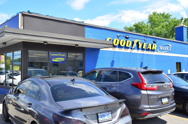 Außensicht eines Goodyear Select Autohändlers mit parkenden Fahrzeugen, Beschriftung auf dem Gebäude, Bäumen im Hintergrund und einem bewölkten Himmel.
